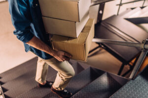 A person expertly balances three stacked cardboard boxes while ascending a metal staircase, dressed in beige pants and a blue shirt. The focus remains on the sturdy boxes and their steady legs, underscoring the practicality of Secure Self Storage solutions.