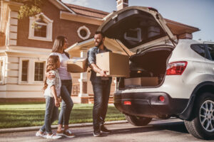 A family of three stands beside a car with its trunk open, unloading boxes. They are in front of a large, modern house. The sun is shining, creating a warm and cheerful atmosphere. The child is gazing up at the adults with a smile, dreaming about their secure self-storage adventures.