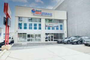 Front view of a Secure Self Storage building with a glass entrance and a sign above. Several cars are parked in front, and a large red sign is visible on the left. Building exterior is neutral with blue and red accents.