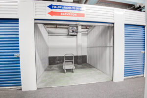 An empty storage unit with a blue roll-up door is open, revealing a dolly inside. A sign above directs to an elevator and the office with blue and red arrows. The unit has corrugated metal walls and a concrete floor.