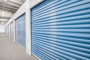 A row of closed blue storage unit doors inside a well-lit warehouse. The floor is carpeted, and the ceiling has exposed beams and overhead lights.