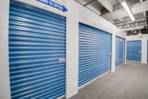 A row of blue, roll-up storage unit doors in a well-lit indoor facility. The ceiling shows exposed beams and fluorescent lighting, with a sign pointing towards the office area.