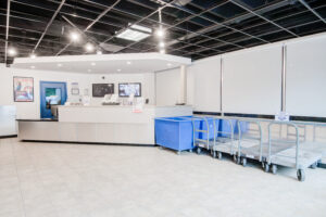 A laundromat interior with a service counter and laundry carts. The ceiling is visibly unfinished with exposed beams, and the floor is tiled. Fluorescent lights are on, and a couple of posters are on the walls.
