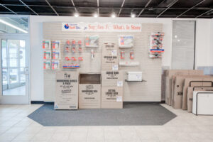 A self-storage store interior displaying various moving supplies. The wall showcases packing items, including boxes, tape, and markers. Large, medium, and wardrobe boxes are neatly stacked on a gray carpeted floor.