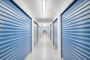 A long corridor of a storage facility with rows of blue roll-up doors on both sides. The floor is white, and the ceiling has fluorescent lights, creating a bright and clean atmosphere.