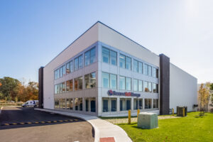 A modern, two-story building with large windows, labeled "Secure Self Storage." The building is surrounded by a paved driveway and green lawns, with a parked truck nearby under clear blue skies.