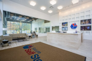 Modern self-storage facility lobby with large windows, a white reception desk, and a beige carpet featuring the "Secure Self Storage" logo. The walls display several televisions, and there is a seating area with gray couches and a coffee table.