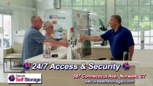 A man in a blue shirt shakes hands with a couple across a counter at a Secure Self Storage facility. A van with the company logo is seen in the background. Text on the image provides contact information and highlights 24/7 access and security.