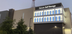 A modern four-story building with illuminated "Secure Self Storage" signage at the top. The structure features large windows and is surrounded by greenery. An antenna extends from the roof against a dusky sky.
