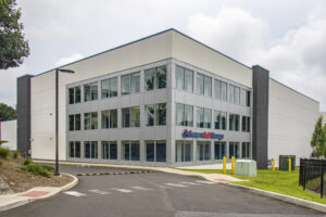 A modern, rectangular two-story building with large windows. The sign "Secure Self Storage" is visible on the facade. The building is surrounded by a paved road and green grass. The sky is cloudy.