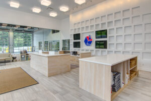 A modern, open lobby with light wood reception desks, a wall-mounted logo, and large windows. The interior features a grid-patterned wall, ceiling lights, and tiled flooring, creating a spacious and welcoming atmosphere.