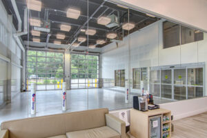 Interior of a well-lit auto service center with large glass windows, reflecting an outdoor view. The space features beige couches, a coffee machine, and a small display shelf in the foreground.