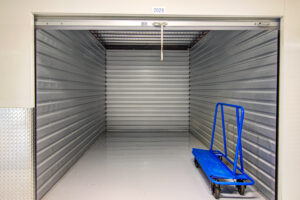 Empty storage unit with open door, revealing clean metal walls and floor. A blue flatbed cart with wheels is positioned to the right inside. The unit has a number "2029" displayed at the top.