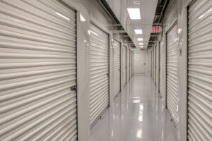A clean indoor storage facility hallway with white roll-up doors on both sides. The floor is shiny and reflective, and there are bright overhead fluorescent lights. An exit sign is visible at the end of the corridor.