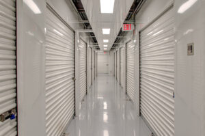 A brightly lit storage facility hallway with shiny white floors and walls. The corridor has rows of closed white metal roll-up storage unit doors on both sides. An illuminated exit sign hangs from the ceiling near the end of the hallway.