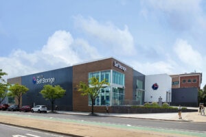 Street view of a modern self-storage facility with large windows and "Secure Self Storage" signage. The building has a mix of dark gray and wooden panels, with a logo on one side. A few trees and parked cars line the street.