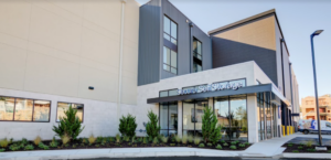 A modern self-storage facility with large windows and a mix of light and dark exterior panels. Lush landscaping with shrubs lines the building's entrance. "Secure Self Storage" is visible above the glass door.