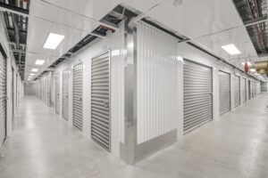 A modern indoor storage facility with metal roll-up doors. The corridor is clean and well-lit, with white walls and a polished concrete floor. The ceiling shows exposed ducts and fixtures. The space appears organized and secure.
