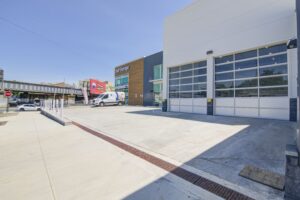 A modern building with glass garage doors and a concrete driveway on a sunny day. There's a white van parked nearby, and an elevated train track in the background. A colorful mural adorns a nearby structure.