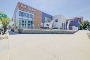A self-storage facility with a modern exterior. A white van is parked next to the building. Two tall feather flags stand near the entrance. A sign displays "Self Storage" on the upper facade, and a contact number is visible. A tree lines the sidewalk.