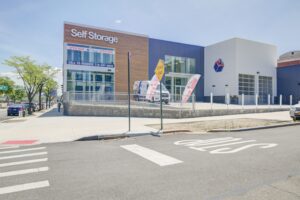 Contemporary self-storage facility with large glass windows and a modern facade. Signs display "Open Now" and a phone number for reservations. The entrance has banners and a visible stop line on the road. Sparse greenery is present.