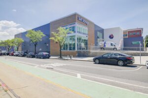 Street view of a building with "Self Storage" signage. The facility features large windows, several trees lined along the sidewalk, a bike lane, and parked cars. A van is seen next to the building under a clear sky.