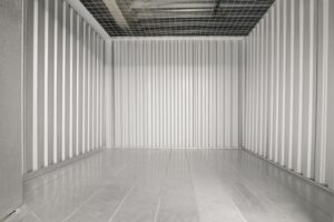An empty metal storage unit with corrugated walls and a wire-mesh ceiling, reflecting light on the shiny floor, creating a stark and industrial appearance.